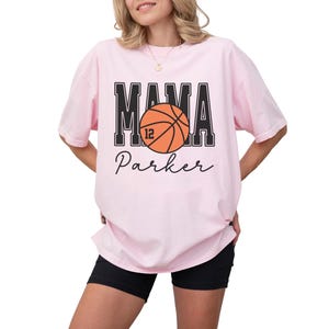 custom basketball mom shirt. the basketball shirt says mama in large varsity letters. there is a large basketball in the middle that is customized with the players number. the basketball tshirt is personalized with the players name below the basketball in a script font. comfort colors basketball mama shirt, short sleeves, crewneck tee