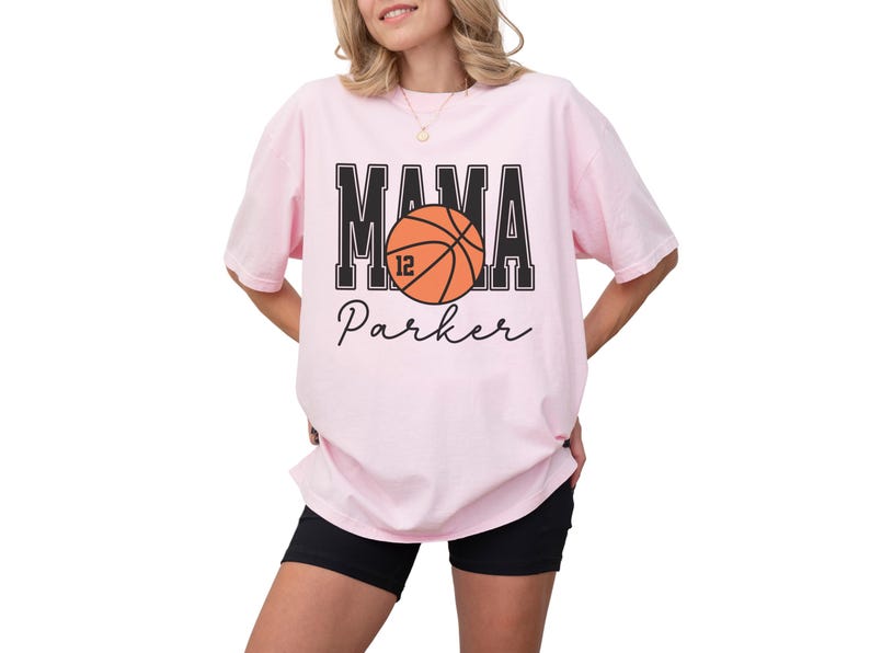 custom basketball mom shirt. the basketball shirt says mama in large varsity letters. there is a large basketball in the middle that is customized with the players number. the basketball tshirt is personalized with the players name below the basketball in a script font. comfort colors basketball mama shirt, short sleeves, crewneck tee