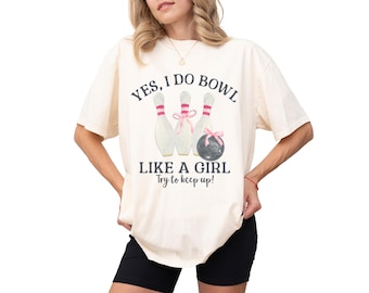May include: Cream-coloured t-shirt with the text "YES, I DO BOWL LIKE A GIRL Try to keep up!" featuring bowling pins and a bowling ball with pink bows. The shirt is a casual style.
