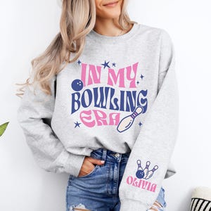 May include: Gray sweatshirt with the text "In My Bowling Era" in pink and blue lettering with a bowling pin and ball graphic. The left sleeve has a bowling pin and ball graphic with the name "Olivia" in pink lettering.