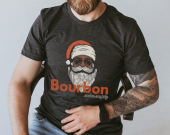 Könnte beinhalten: Dunkelgraues T-Shirt mit einer Santa Claus-Grafik mit Sonnenbrille und weißem Bart. Der Text "Bourbon makes me jolly" ist darunter in Orange und Weiß aufgedruckt. Das T-Shirt hat kurze Ärmel.