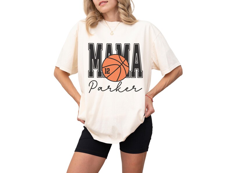 custom basketball mom shirt. the basketball shirt says mama in large varsity letters. there is a large basketball in the middle that is customized with the players number. the basketball tshirt is personalized with the players name below the basketball in a script font. comfort colors basketball mama shirt, short sleeves, crewneck tee