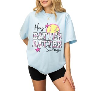 May include: Light blue t-shirt with the text "Hey Batter Batter Swing" in black and pink. The design includes a yellow softball, pink stars, and hearts. The shirt is a casual, oversized fit.
