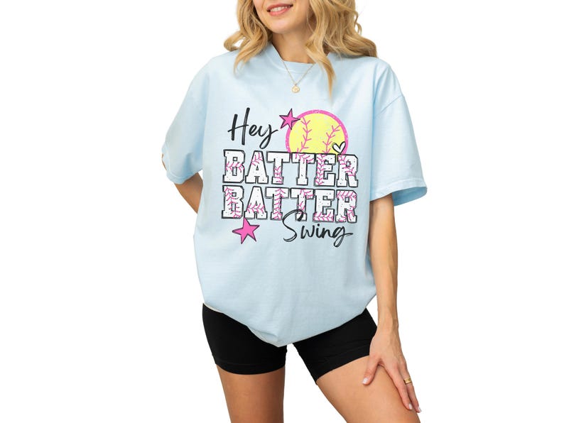 May include: Light blue t-shirt with the text "Hey Batter Batter Swing" in black and pink. The design includes a yellow softball, pink stars, and hearts. The shirt is a casual, oversized fit.
