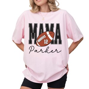 Custom Football Mama Shirt. The tshirt says football mama in varsity font and is personalized with the players number on a large football and name below it. Comfort Colors tee, crewneck, short sleeves football mom t shirt
