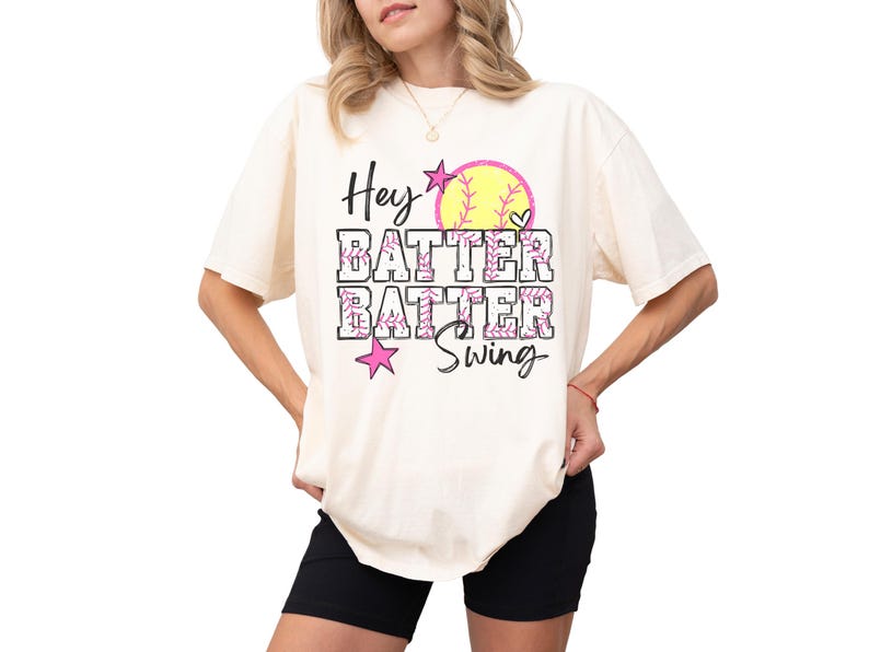 May include: Cream-colored t-shirt with the text "Hey Batter Batter Swing" in black, pink, and yellow. The design includes a softball and stars. The shirt is a casual, oversized fit.