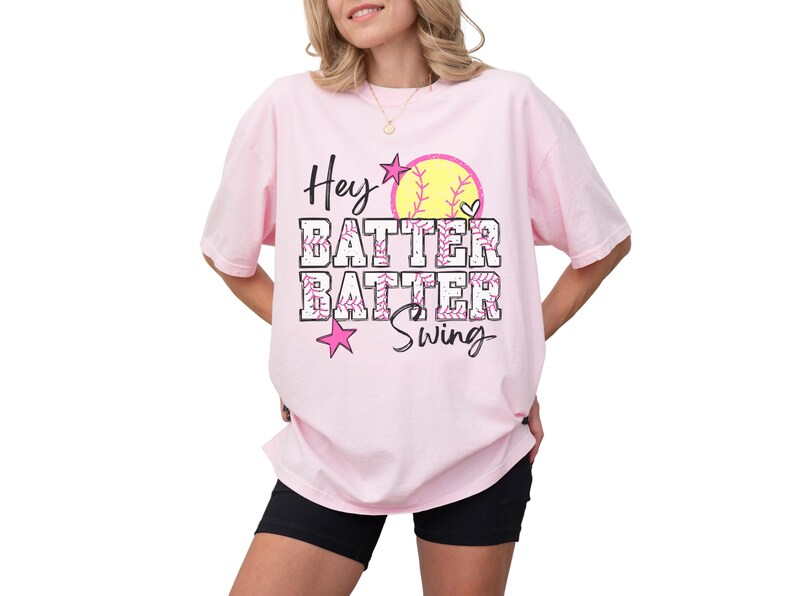 May include: A light pink t-shirt with the words "Hey Batter Batter Swing" in black and pink. The design includes a yellow softball with a heart and pink stars. The shirt is worn with black shorts.