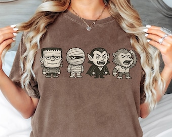 Pode incluir: Camiseta marrom com quatro personagens de desenho animado de Halloween: o monstro de Frankenstein, uma múmia, um vampiro e um lobisomem. Os personagens estão em fila, cada um com detalhes e expressões únicas. A camiseta é marrom lisa.