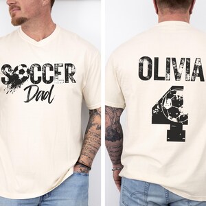 May include: A white t-shirt with a black graphic that says "Soccer Dad" with a soccer ball. The back of the shirt has a black graphic with the name "Olivia" and the number "4" with a soccer ball.