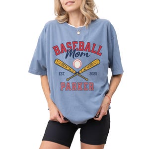 baseball mom shirt. the baseball mom tee says baseball mom and is personalized with the player name and the year. The graphic has two baseball bats and a baseball. comfort colors, short sleeves, crewneck, baseball tshirt for mama