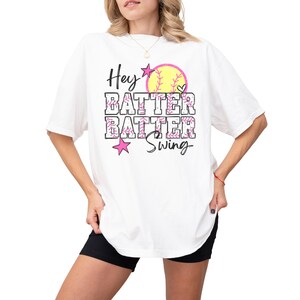 May include: White t-shirt with the text "Hey Batter Batter Swing" in black and pink. A yellow softball with pink stitching and a heart graphic is above the text. The shirt is a casual style.
