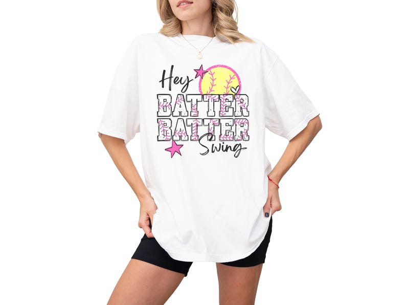 May include: White t-shirt with the text "Hey Batter Batter Swing" in black and pink. A yellow softball with pink stitching and a heart graphic is above the text. The shirt is a casual style.