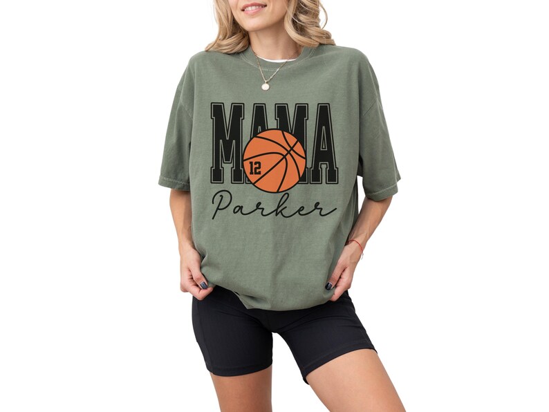 custom basketball mom shirt. the basketball shirt says mama in large varsity letters. there is a large basketball in the middle that is customized with the players number. the basketball tshirt is personalized with the players name below the basketball in a script font. comfort colors basketball mama shirt, short sleeves, crewneck tee