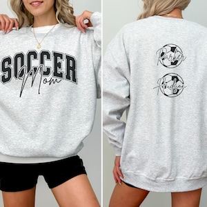 May include: Light gray sweatshirt with the text "SOCCER Mom" in black and white lettering on the front. The back of the sweatshirt has two soccer ball designs with the names "Parker" and "Hudson" in black script.