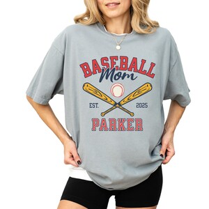 baseball mom shirt. the baseball mom tee says baseball mom and is personalized with the player name and the year. The graphic has two baseball bats and a baseball. comfort colors, short sleeves, crewneck, baseball tshirt for mama