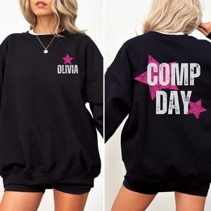 May include: Black sweatshirt with a pink star and the text "Olivia" on the front and a pink star with the text "COMP DAY" on the back.