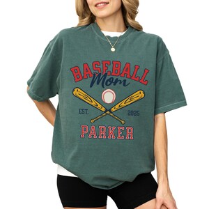baseball mom shirt. the baseball mom tee says baseball mom and is personalized with the player name and the year. The graphic has two baseball bats and a baseball. comfort colors, short sleeves, crewneck, baseball tshirt for mama