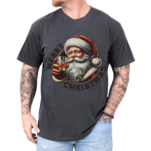 Christmas drinking shirt graphic tee. The t shirt has a distressed, vintage Santa that is holding a glass of whiskey or bourbon on the rocks. The tshirt says Merry Christmas. Comfort Colors tee, short sleeves, crewneck
