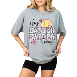 May include: A light gray t-shirt with the words "Hey Batter Batter Swing" in a playful font. The design includes a yellow softball, pink stars, and a heart. The shirt is a comfortable, oversized fit.