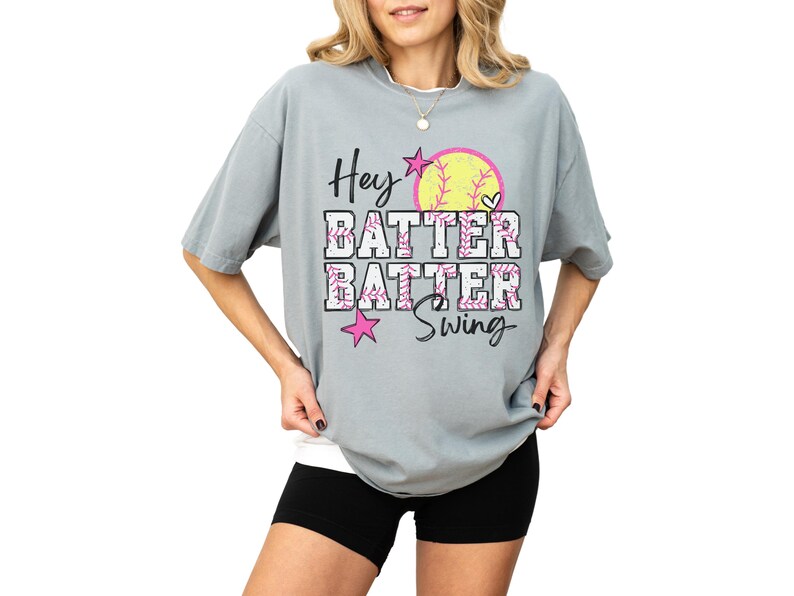 May include: A light gray t-shirt with the words "Hey Batter Batter Swing" in a playful font. The design includes a yellow softball, pink stars, and a heart. The shirt is a comfortable, oversized fit.