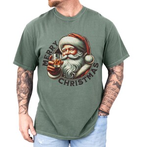 Christmas drinking shirt graphic tee. The t shirt has a distressed, vintage Santa that is holding a glass of whiskey or bourbon on the rocks. The tshirt says Merry Christmas. Comfort Colors tee, short sleeves, crewneck