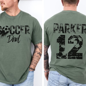 Custom Soccer Shirt for Dad. The soccer tshirt says Soccer Dad on the front in a black varsity collegiate style font. The back is personalized with the players name and number. The number is a collegiate font with a soccer ball pattern. comfort colors tee, short sleeves, crewneck soccer dad tshirt