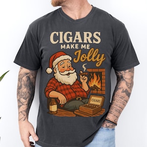 May include: Dark gray t-shirt with a graphic of Santa Claus smoking a cigar in front of a fireplace. The text reads "CIGARS make me Jolly". Santa is wearing a red and black plaid shirt and a Santa hat. A box of cigars and a bottle are also visible.