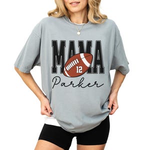 Custom Football Mama Shirt. The tshirt says football mama in varsity font and is personalized with the players number on a large football and name below it. Comfort Colors tee, crewneck, short sleeves football mom t shirt