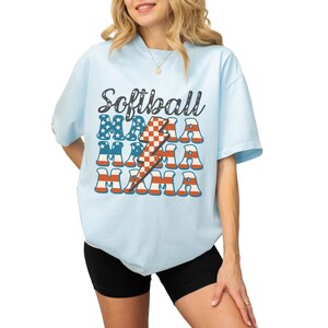 Retro patriotic softball mama shirt for 4th of July and Memorial day. The t shirt says, softball in black distressed script and the word mama repeated 3 times in a red white and blue USA flag font. A checkered lightning bolt down the middle