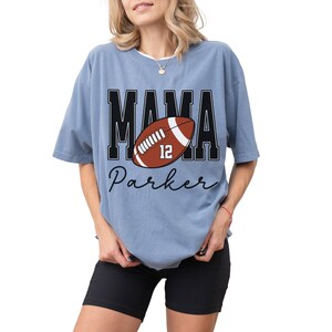 Custom Football Mama Shirt. The tshirt says football mama in varsity font and is personalized with the players number on a large football and name below it. Comfort Colors tee, crewneck, short sleeves football mom t shirt