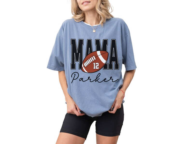 Custom Football Mama Shirt. The tshirt says football mama in varsity font and is personalized with the players number on a large football and name below it. Comfort Colors tee, crewneck, short sleeves football mom t shirt
