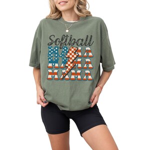 Retro patriotic softball mama shirt for 4th of July and Memorial day. The t shirt says, softball in black distressed script and the word mama repeated 3 times in a red white and blue USA flag font. A checkered lightning bolt down the middle