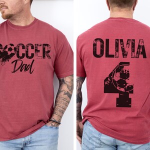 May include: Two red t-shirts with black text and graphics. The first shirt says "Soccer Dad" with a soccer ball graphic. The second shirt says "Olivia" with a soccer ball graphic and the number "4".