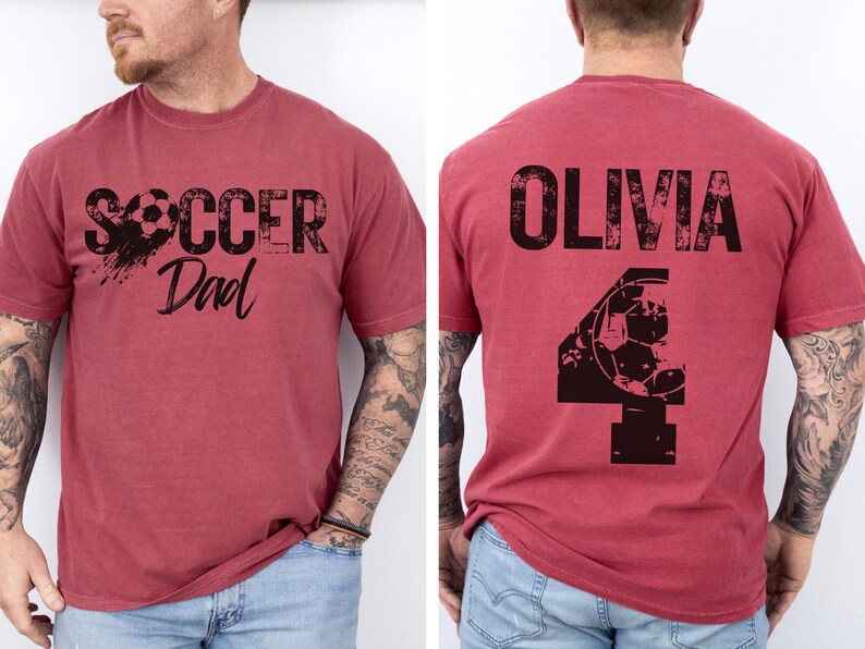 May include: Two red t-shirts with black text and graphics. The first shirt says "Soccer Dad" with a soccer ball graphic. The second shirt says "Olivia" with a soccer ball graphic and the number "4".