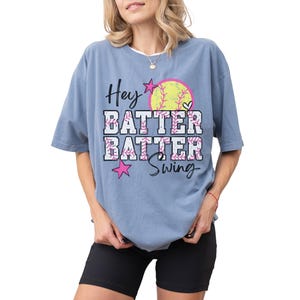 May include: A blue t-shirt with the words "Hey Batter Batter Swing" in a playful font. The design includes a softball, stars, and floral accents. The shirt is a comfortable, oversized fit.