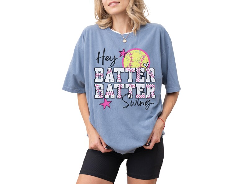 May include: A blue t-shirt with the words "Hey Batter Batter Swing" in a playful font. The design includes a softball, stars, and floral accents. The shirt is a comfortable, oversized fit.