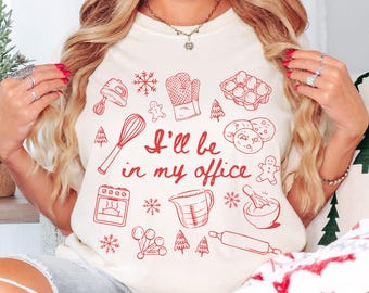 May include: Cream-coloured t-shirt featuring a red graphic of baking tools and the text "I'll be in my office." The design includes a whisk, oven, measuring jug, rolling pin, and gingerbread men. The shirt is a casual, comfortable style.