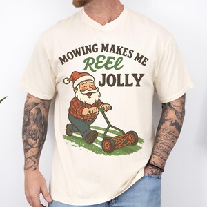 May include: Cream-colored t-shirt with a graphic of Santa Claus mowing grass, wearing a Santa hat and plaid shirt. The text reads "Mowing Makes Me Reel Jolly."