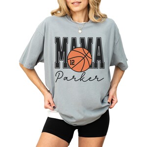 custom basketball mom shirt. the basketball shirt says mama in large varsity letters. there is a large basketball in the middle that is customized with the players number. the basketball tshirt is personalized with the players name below the basketball in a script font. comfort colors basketball mama shirt, short sleeves, crewneck tee