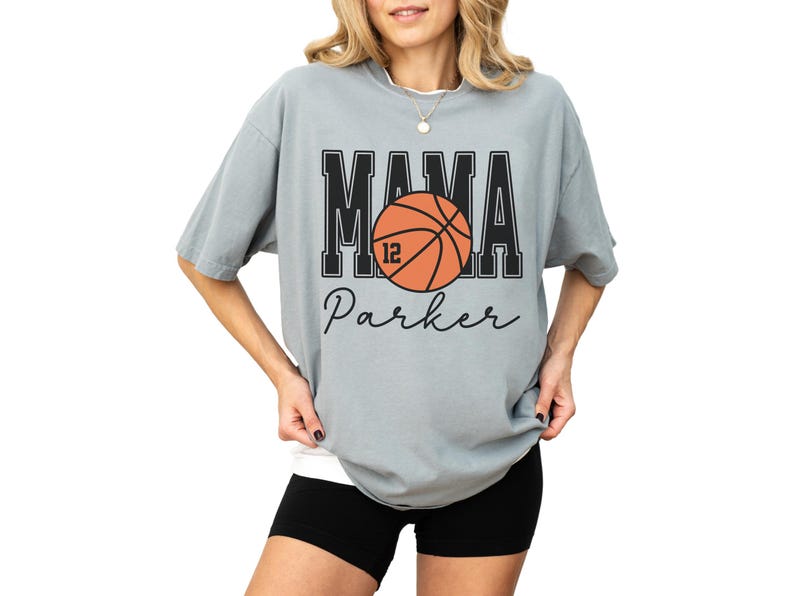 custom basketball mom shirt. the basketball shirt says mama in large varsity letters. there is a large basketball in the middle that is customized with the players number. the basketball tshirt is personalized with the players name below the basketball in a script font. comfort colors basketball mama shirt, short sleeves, crewneck tee