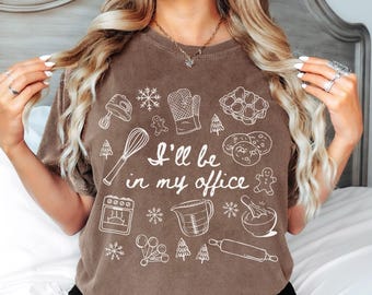 May include: Brown t-shirt featuring white illustrations of baking tools and treats. The text "I'll be in my office" is printed below the illustrations. The design includes a whisk, oven, gingerbread man, and rolling pin.