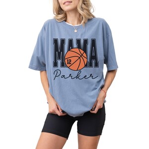 custom basketball mom shirt. the basketball shirt says mama in large varsity letters. there is a large basketball in the middle that is customized with the players number. the basketball tshirt is personalized with the players name below the basketball in a script font. comfort colors basketball mama shirt, short sleeves, crewneck tee