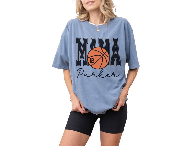 custom basketball mom shirt. the basketball shirt says mama in large varsity letters. there is a large basketball in the middle that is customized with the players number. the basketball tshirt is personalized with the players name below the basketball in a script font. comfort colors basketball mama shirt, short sleeves, crewneck tee