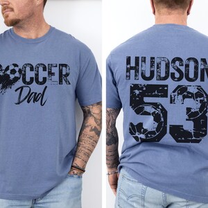 May include: A blue t-shirt with a distressed black graphic that says "Soccer Dad" on the front and "HUDSON 53" on the back.