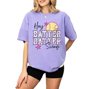 May include: A lilac-colored t-shirt with the words "Hey Batter Batter Swing" in a playful font. A softball graphic and pink stars accent the design. The shirt is a comfortable, oversized fit.