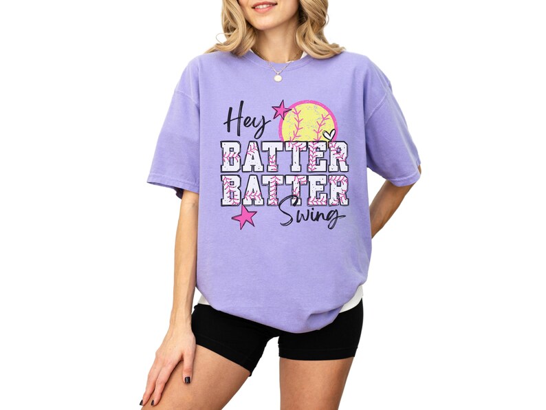 May include: A lilac-colored t-shirt with the words "Hey Batter Batter Swing" in a playful font. A softball graphic and pink stars accent the design. The shirt is a comfortable, oversized fit.