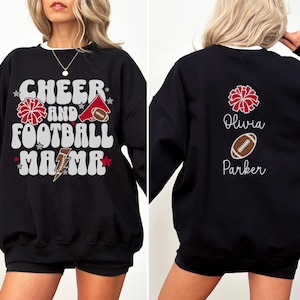 Puede incluir: Sudadera negra con ribete blanco. La parte delantera muestra "Cheer and Football Mama" con un diseño de pompón rojo, fútbol americano y megáfono. La parte trasera presenta un pompón rojo, un balón de fútbol y los nombres "Olivia Parker".