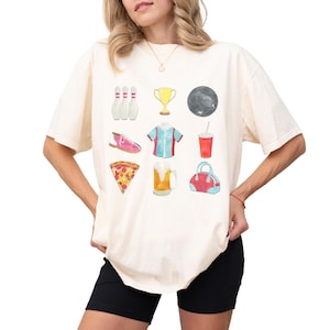 Retro bowling shirt for women. The tshirt has watercolor graphics and features bowling pins, trophy, bowling ball, shoes, shirt, soda, pizza, beer and a bowling bag. Short sleeves, crewneck, comfort colors, ivory
