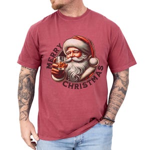Christmas drinking shirt graphic tee. The t shirt has a distressed, vintage Santa that is holding a glass of whiskey or bourbon on the rocks. The tshirt says Merry Christmas. Comfort Colors tee, short sleeves, crewneck