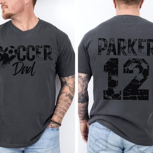 May include: A dark gray t-shirt with the text "Soccer Dad" and a soccer ball graphic on the front. The back of the shirt has the text "Parker 12" with a soccer ball graphic.